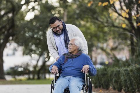 Man wearing glasses in sweater and scarf pushing older man wearing glasses in wheelchair wearing scarf