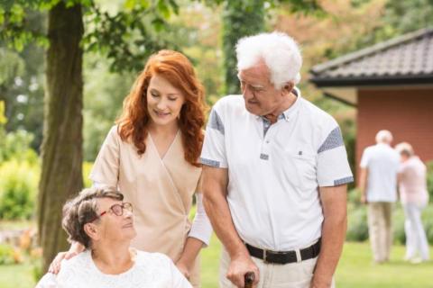 Older woman in wheelchair being pushed by young woman visiting with older man with cane in outside setting