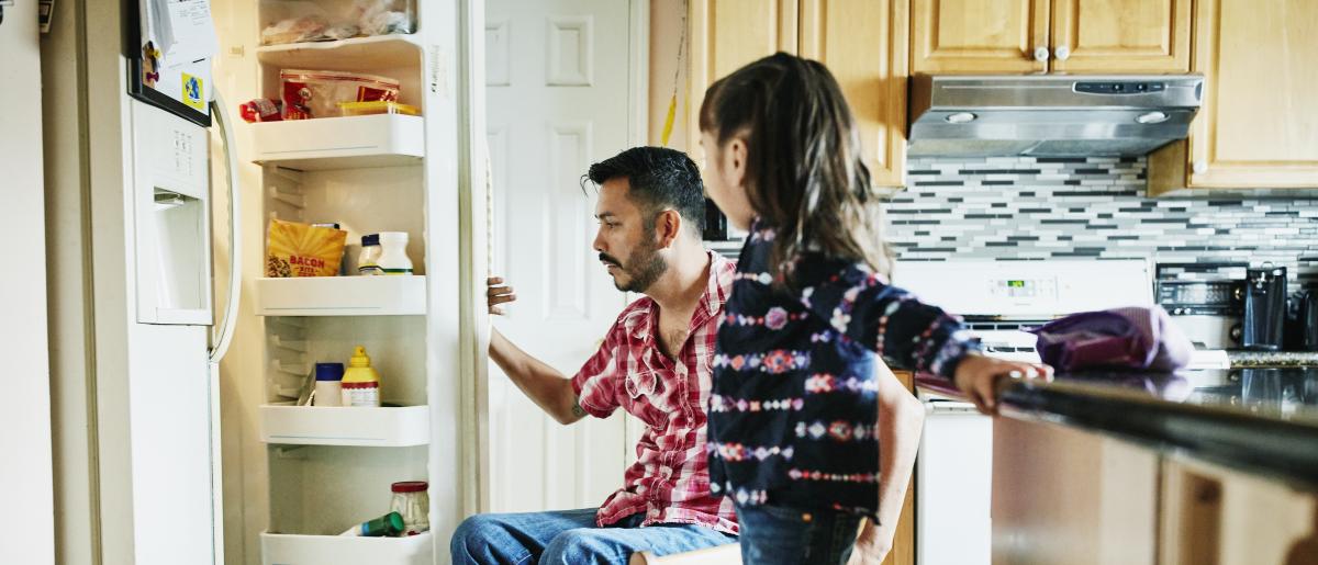 Man in wheelchair and young girl standing on chair looking inside opened refrigerator in kitchen