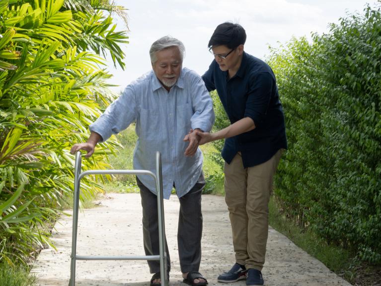 Younger man wearing glasses assisting older man with walker in a garden setting