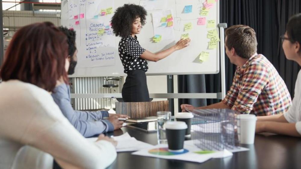 African American woman in front of a board presenting to a group of colleagues sitting at a conference table