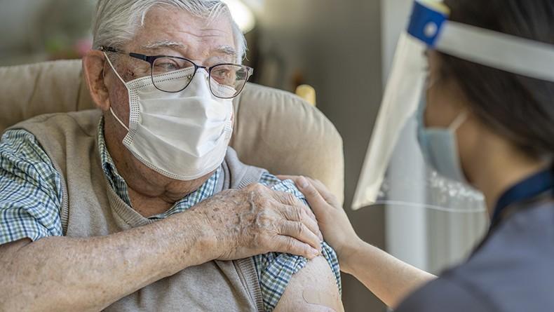 Older gentleman wearing mask and glasses in chair holding sleeve talking with nurse wearing face shield and mask following vaccine administration