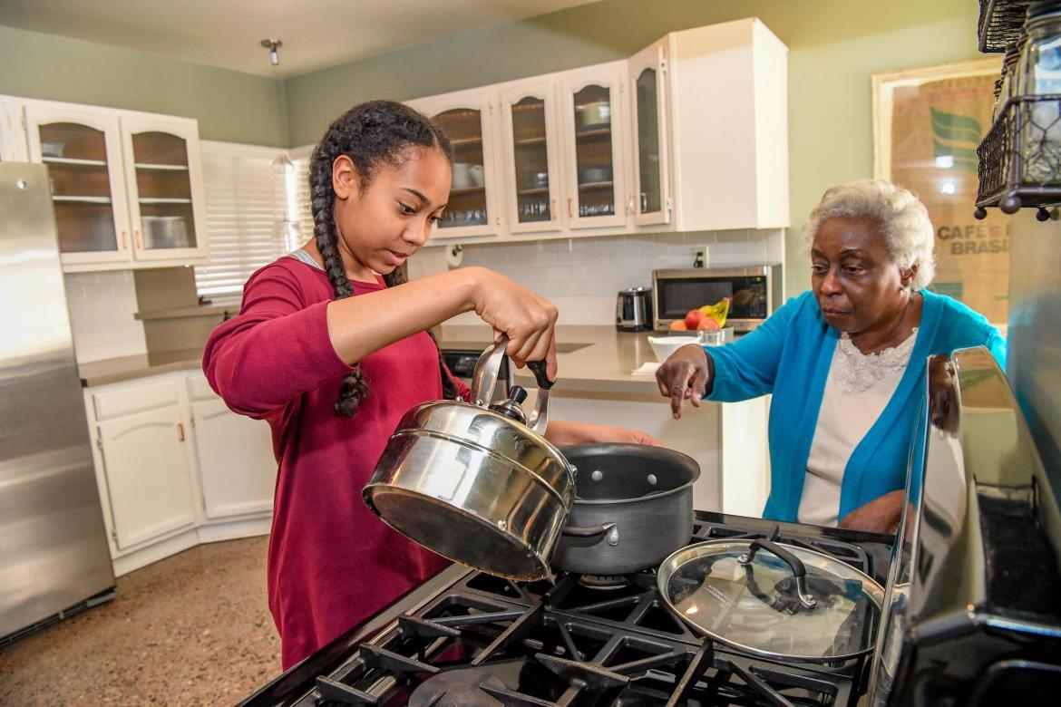 Young girl being instructed by grandmother pouring water from a teakettle into an empty pot on the stove in a kitchen